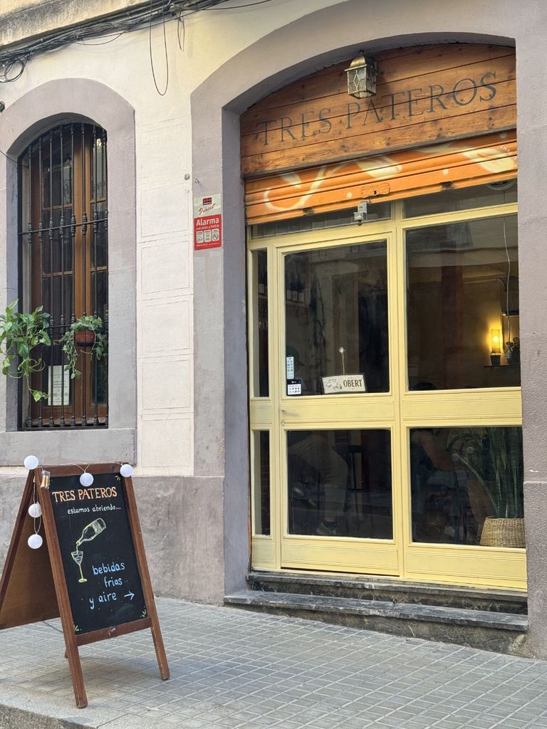 Façana de Tres Pateros: el rètol de fusta amb el nom sobre la porta de vidre groga i una pissarra a la vorera