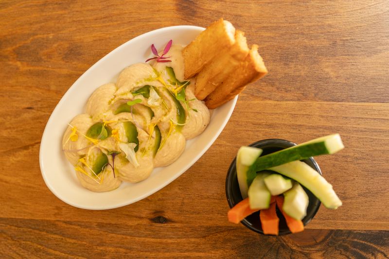 Hummus visto desde arriba con pan tostado, brotes y crudités de pepino y zanahoria sobre una mesa de madera