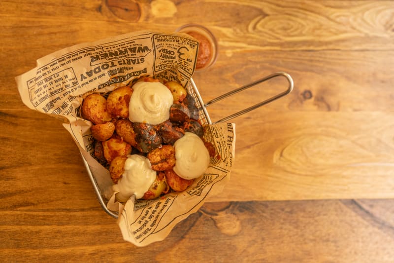 Patatas pateras vistas desde arriba: patatas especiadas con trozos de embutido y mayonesa de ajo en una cesta de alambre con papel de periódico, junto a un cuenco de salsa roja sobre una mesa de madera