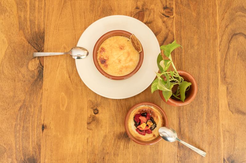 Vista cenital de dos postres en cazuelas de barro, uno con frutos rojos y flores comestibles, sobre una mesa de madera junto a una planta